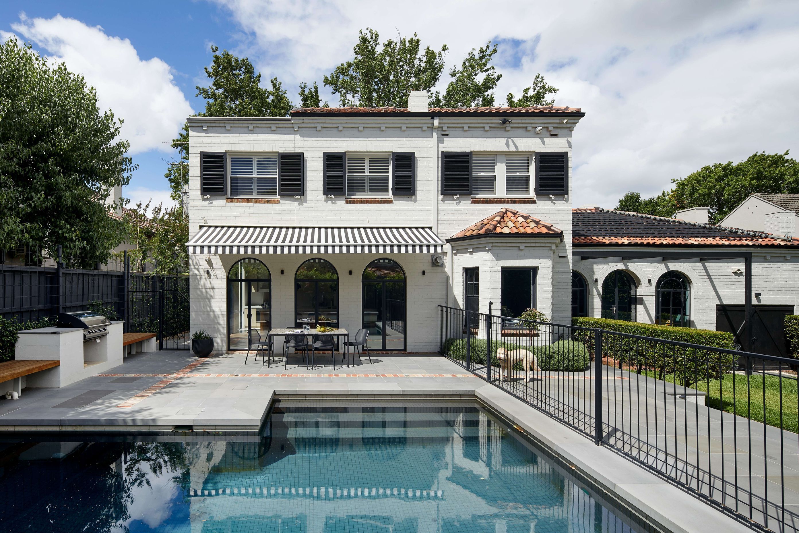 Art deco spanish mission house from the 1930’s renovated with updated paint with charcoal shutters. Rear space off kitchen features black and white striped awning, from Docril. See more from our Arch Deco Project.