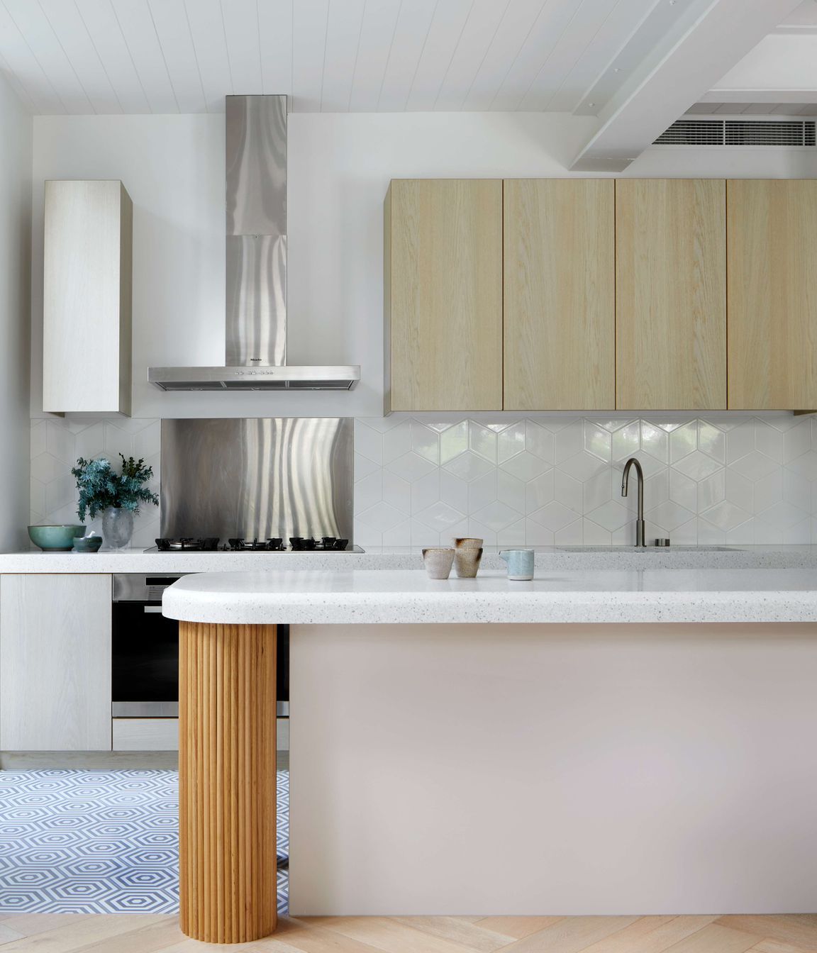 White kitchen with minimalist styling featuring white gloss diamond Perini tile backsplash, vibrant oak cool white veneer cabinetry, and a central cool grey cement hexagon patterned tiles on the floor, silver birch coloured corian stone bench top and oversized hand crafted pedestal legs using half round dowels as the finish for a contemporary and modern feel. Located in Melbourne, see more from our Arch Deco Project.