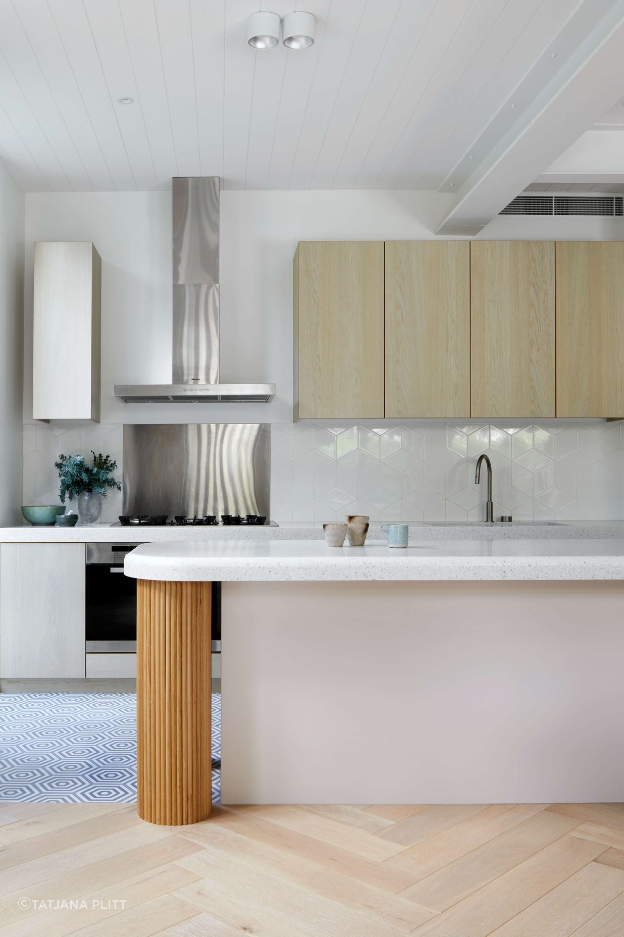 White kitchen with minimalist styling featuring white gloss diamond Perini tile backsplash, vibrant oak cool white veneer cabinetry, and a central cool grey cement hexagon patterned tiles on the floor, silver birch coloured corian stone bench top and oversized hand crafted pedestal legs using half round dowels as the finish for a contemporary and modern feel. Located in Melbourne, see more from our Arch Deco Project.