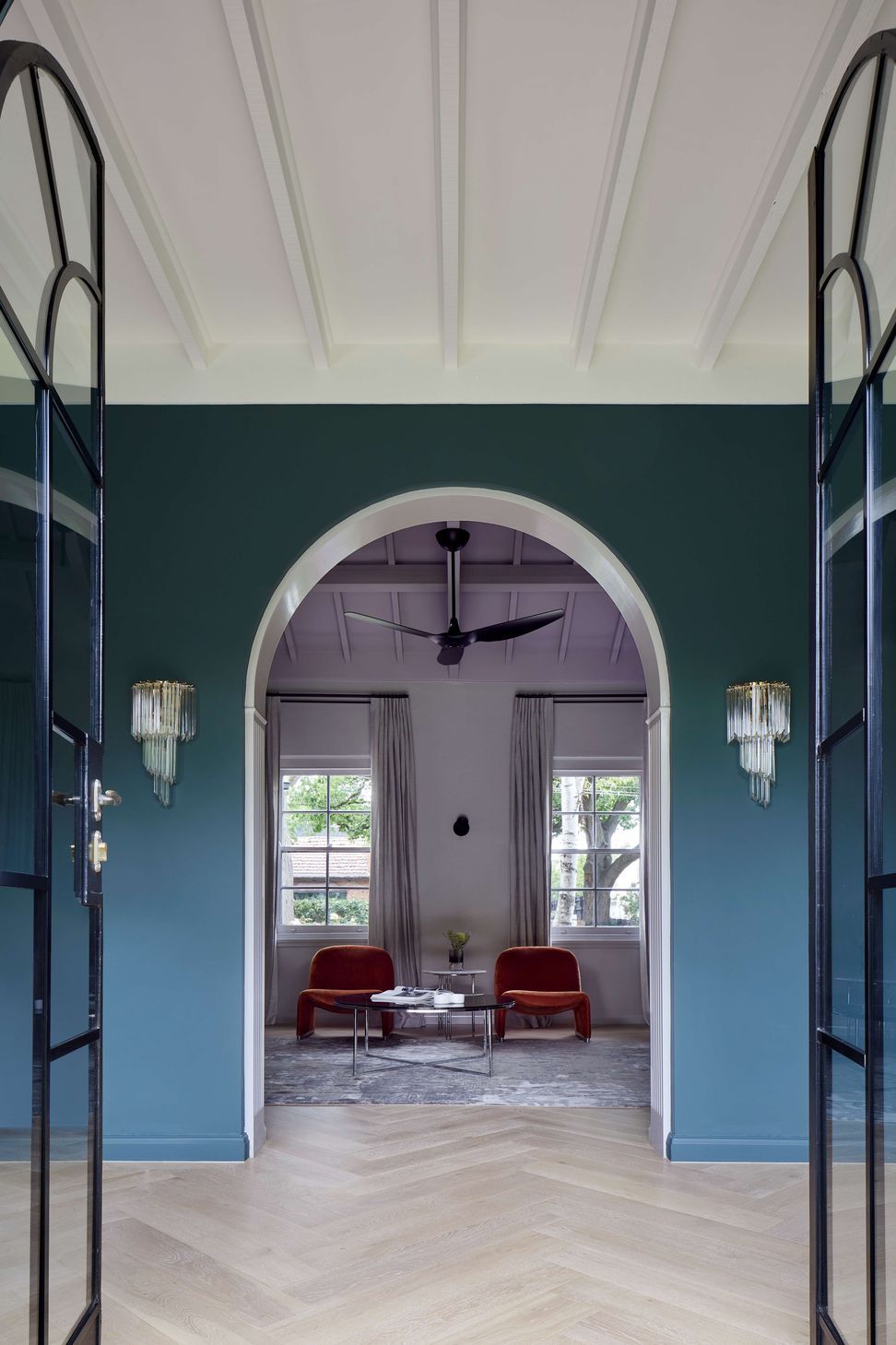 Library view from the living room through arched doorways and ceiling beams to keep with the art deco style of this house. Wall painted in dark green (Coriole) from Dulux (colour: Coriole). Crystal wall pendant lights frame the doorway and flooring is european oak laid in herringbone pattern with a matt lacquer for a minimalist styling.Project is a 1930s art deco spanish mission style house in Melbourne. See more from our Arch Deco Project.