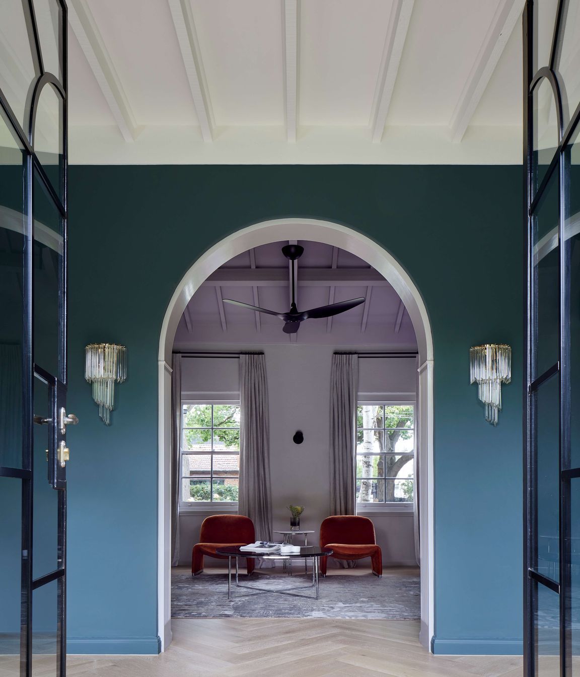Library view from the living room through arched doorways and ceiling beams to keep with the art deco style of this house. Wall painted in dark green (Coriole) from Dulux (colour: Coriole). Crystal wall pendant lights frame the doorway and flooring is european oak laid in herringbone pattern with a matt lacquer for a minimalist styling.Project is a 1930s art deco spanish mission style house in Melbourne. See more from our Arch Deco Project.