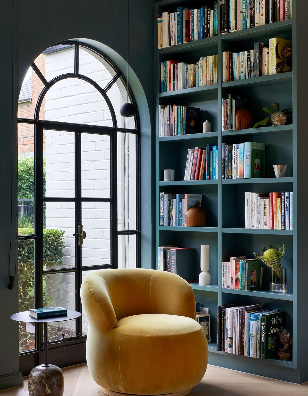 Art deco library featuring a large arched doorway framed by bookcases. Wall and bookcase painted in dark green from Dulux (colour: Coriole). Occasional Joy chair upholstered in elk velvet mustard from Jardan. Flooring in oak wood laid in herringbone pattern with a matt lacquer for a minimalist styling. The project is a 1930s art deco spanish mission style house in Melbourne. See more from our Arch Deco Project.