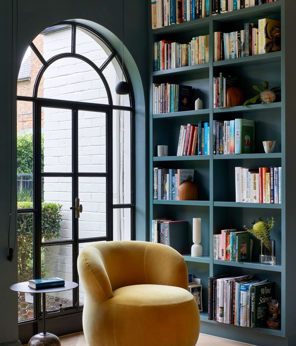 Art deco library featuring a large arched doorway framed by bookcases. Wall and bookcase painted in dark green from Dulux (colour: Coriole). Occasional Joy chair upholstered in elk velvet mustard from Jardan. Flooring in oak wood laid in herringbone pattern with a matt lacquer for a minimalist styling. The project is a 1930s art deco spanish mission style house in Melbourne. See more from our Arch Deco Project.