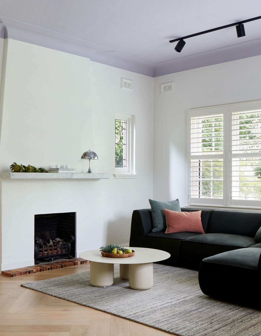 TV room featuring 1930s art deco fireplace, with marble mantle and a Louis Pulsen Panthella light from Cult. The flooring is oak wood herringbone, coffee table from Coco Flip. Emerald upholstered sofa from Jardan adds a pop of colour to a simply styled white TV room with mauve ceiling. See more from our Arch Deco Project.