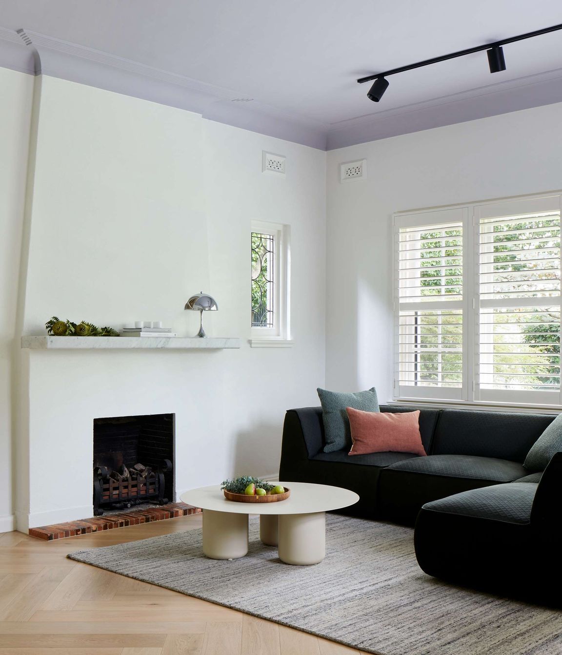 TV room featuring 1930s art deco fireplace, with marble mantle and a Louis Pulsen Panthella light from Cult. The flooring is oak wood herringbone, coffee table from Coco Flip. Emerald upholstered sofa from Jardan adds a pop of colour to a simply styled white TV room with mauve ceiling. See more from our Arch Deco Project.