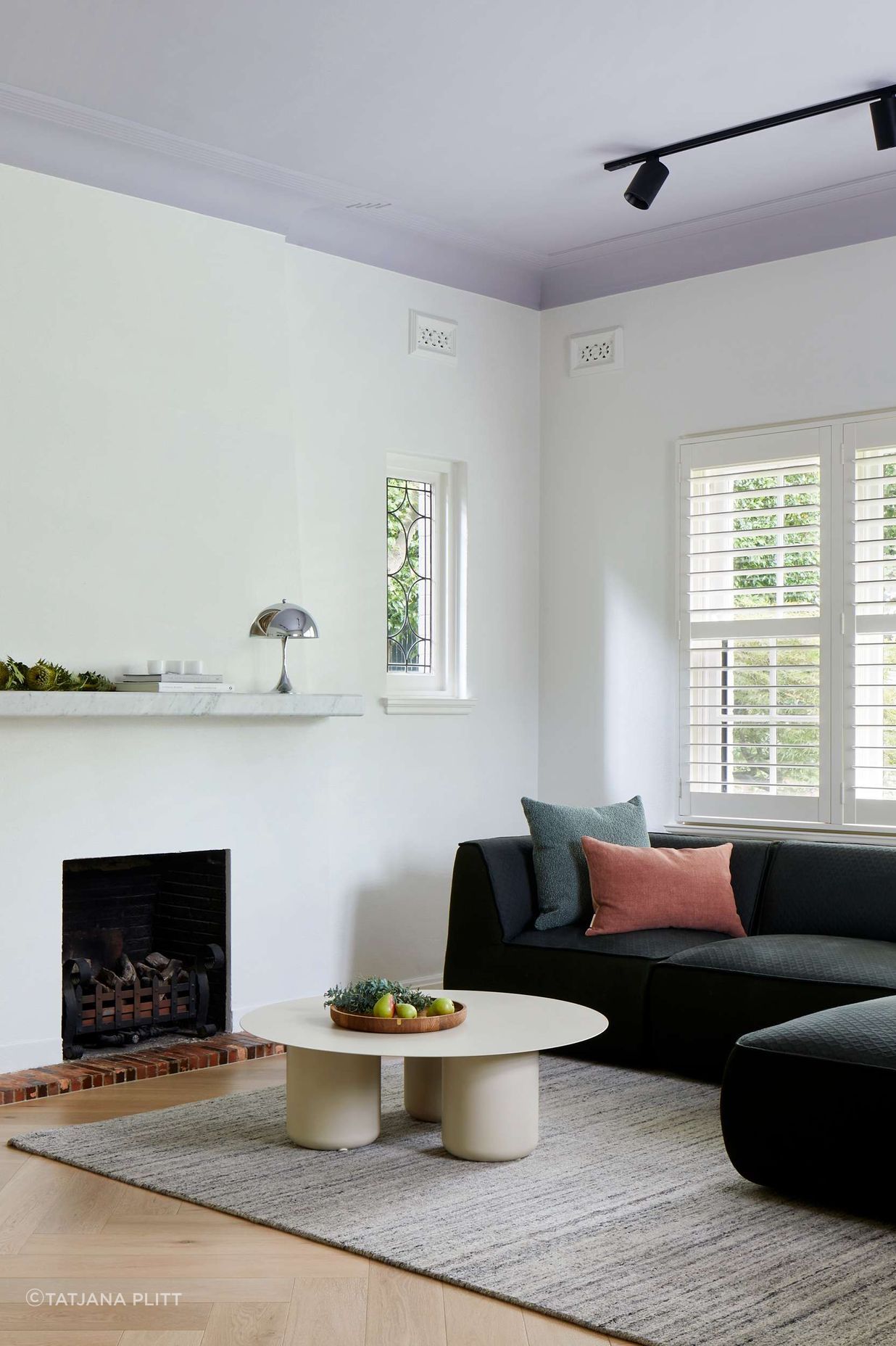 TV room featuring 1930s art deco fireplace, with marble mantle and a Louis Pulsen Panthella light from Cult. The flooring is oak wood herringbone, coffee table from Coco Flip. Emerald upholstered sofa from Jardan adds a pop of colour to a simply styled white TV room with mauve ceiling. See more from our Arch Deco Project.