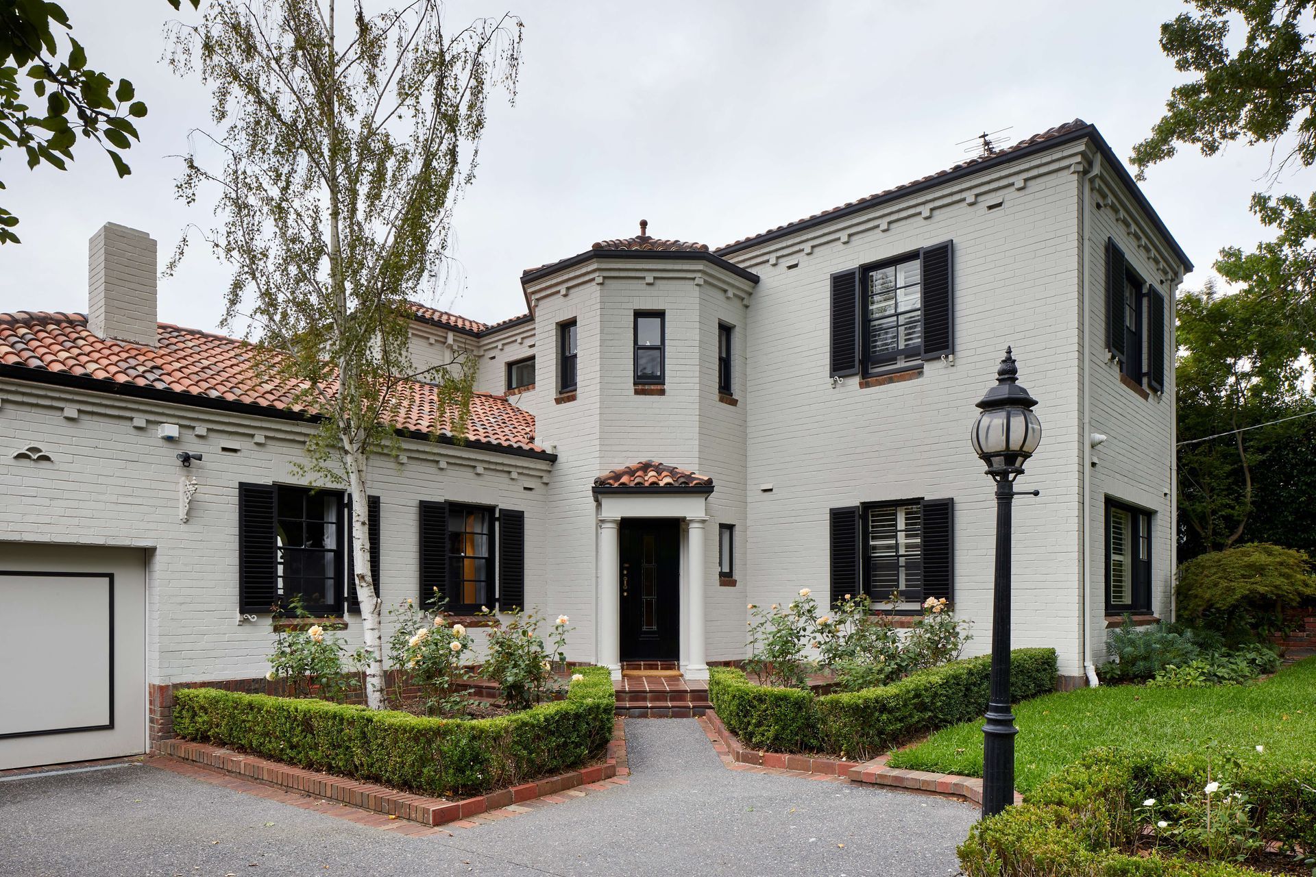 Renovated 1930’s art deco spanish mission style house in Melbourne. Art deco spanish mission house from the 1930’s renovated with updated paint with black shutters. Front of house with formal garden with box hedges and terracotta tiles. See more from our Arch Deco Project.