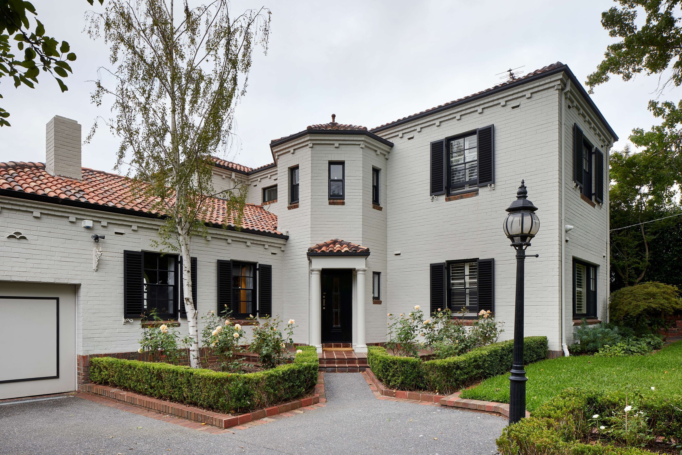 Renovated 1930’s art deco spanish mission style house in Melbourne. Art deco spanish mission house from the 1930’s renovated with updated paint with black shutters. Front of house with formal garden with box hedges and terracotta tiles. See more from our Arch Deco Project.