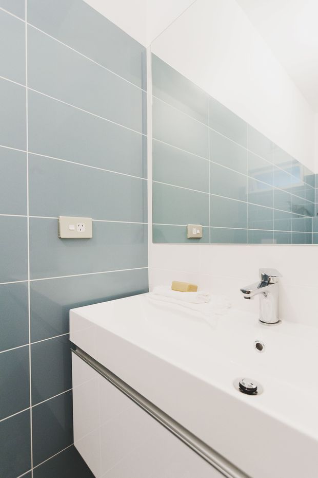Bright, contemporary bathroom vanity area featuring a white integrated basin and chrome tapware, white built-in cabinetry, pale blue-grey large-format wall tiles with white grout, and a large wall mirror.
