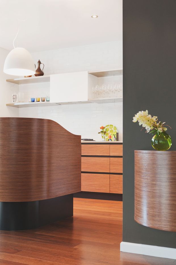 A contemporary kitchen viewed through a dark charcoal partition, featuring warm timber flooring, a curved dark wood island or breakfast bar, matching timber cabinetry, white subway-tile walls, open shelving, and a large white pendant light. A small timber-clad pedestal with a vase adds a decorative foreground element.