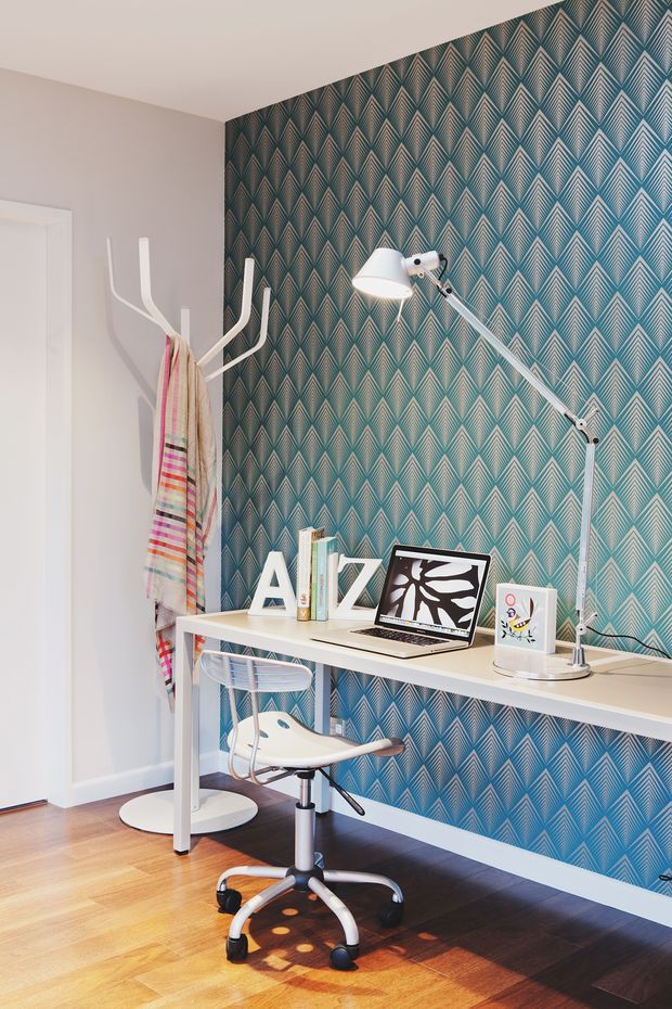 Compact, brightly lit home office with a white desk against a bold teal and grey geometric patterned feature wall. The workspace includes a laptop, articulated desk lamp, books, framed artwork, and a modern white office chair. A sculptural coat stand with a colourful textile sits beside the desk, while timber flooring and pale walls complete the room.