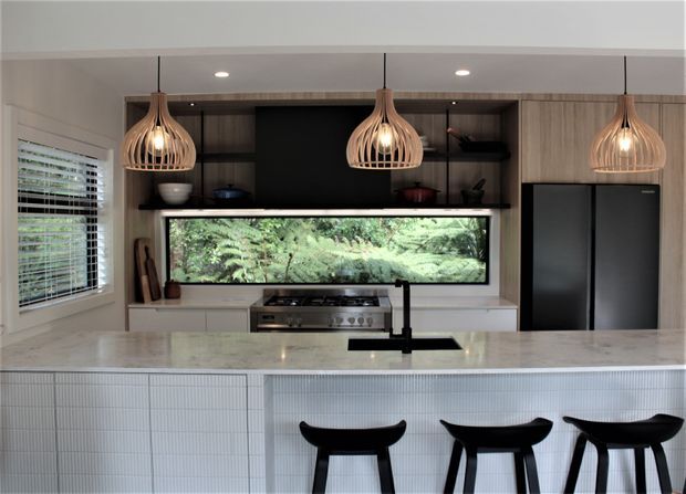 Contemporary kitchen with a large white island, integrated gas cooktop and black gooseneck tap, full-height timber-look cabinetry, matte black refrigerator, open display shelving, and three sculptural pendant lights. A long illuminated fern-filled feature window or indoor planter sits behind the cooktop, while black bar stools line the island. The palette is light and neutral with warm timber accents and strong black contrast.
