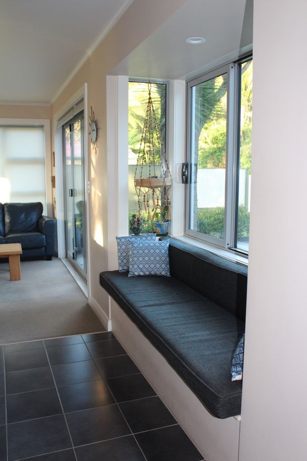 A bright, narrow residential sunroom or sitting area with a built-in window seat running along a wall of large windows. The room includes dark upholstered seating with patterned cushions, black square floor tiles in the foreground, pale walls and ceiling, and a carpeted adjoining lounge area with a leather armchair. Indoor plants and hanging decorative elements are visible by the glazing, with greenery outside.