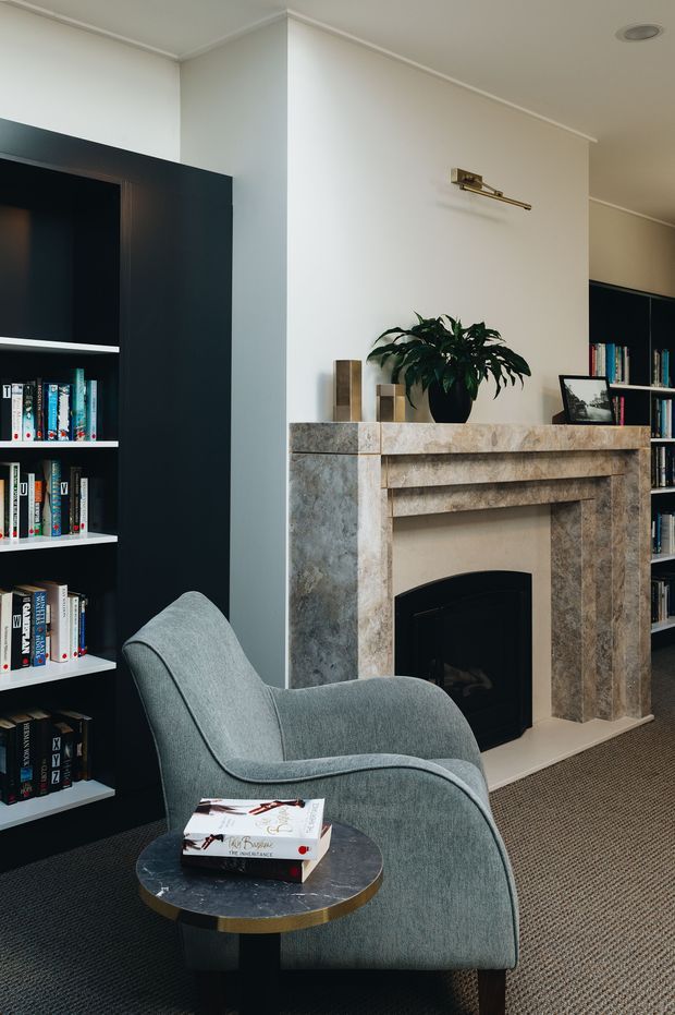 A refined living room with a built-in dark bookcase, upholstered lounge chair, round side table, and a stone-clad fireplace with a brass picture light above. The room has pale walls, dark carpet, curated books, and understated contemporary-traditional detailing.