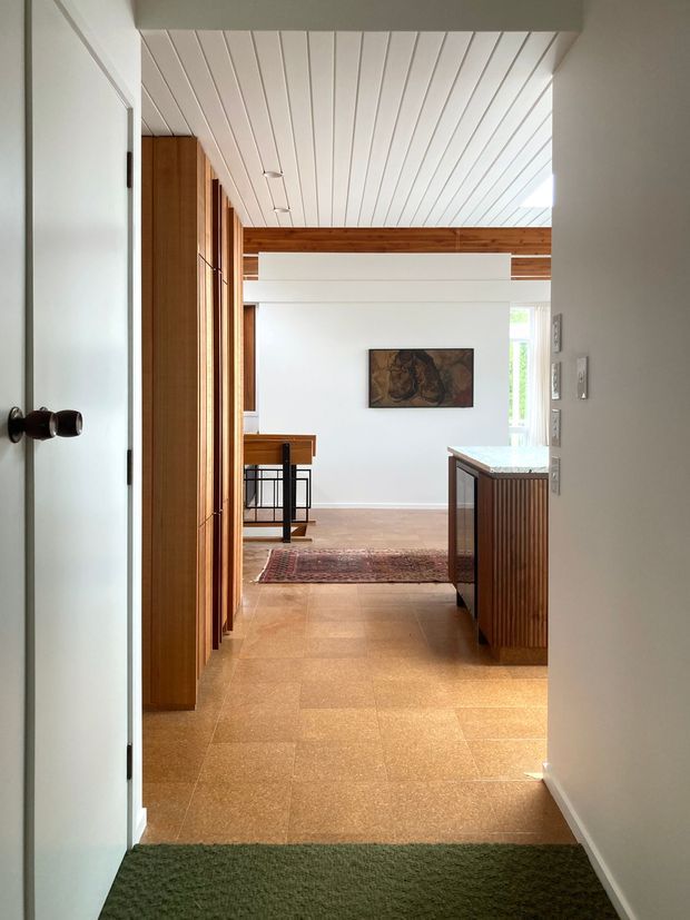 View through an interior doorway into a bright, open-plan residential space. The foreground has a white door and dark green carpet, opening onto warm tan tiled flooring. Vertical timber cabinetry or wall panelling lines the left side, while a timber-slatted island or built-in unit with a pale stone benchtop is on the right. The room beyond has white walls, a white-lined ceiling with exposed timber beams, a small dining or kitchen area, a rug, and artwork. The design combines warm timber detailing with clean white surfaces and natural light.