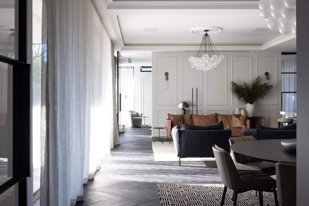 A refined open-plan living and dining room with white decorative wall panelling, coffered ceiling detailing, full-height sheer curtains, dark herringbone timber flooring, and contemporary furniture. The seating area includes a tan sofa, dark textured armchairs, and a sculptural clustered pendant light, while a dark dining table and upholstered dining chairs occupy the foreground. Large glazing along the left side fills the space with natural light.