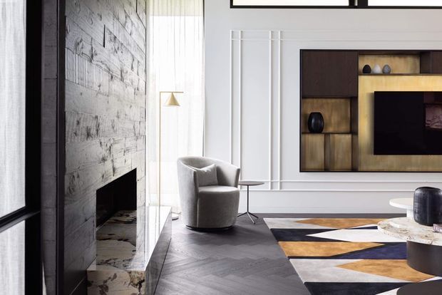 Refined contemporary living room with a dramatic dark stone fireplace wall, herringbone timber flooring, white paneled walls, and a built-in dark timber and brass-toned media unit. The room includes a sculptural upholstered lounge chair, side table, patterned geometric rug, and soft natural light through tall sheer curtains.