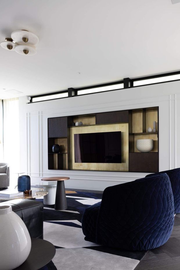 A refined living room featuring a large built-in TV and display wall with dark timber cabinetry, warm metallic-toned backing, and white decorative wall panelling. The room includes sculptural lounge chairs and sofas in white, grey, and deep navy, a geometric monochrome rug, dark timber flooring, and contemporary decorative lighting. Black-framed clerestory glazing runs above the feature wall, bringing in natural light.