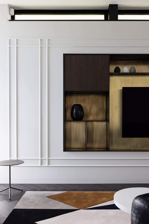 Contemporary living room interior featuring a sculptural built-in dark timber entertainment and display unit set into a white paneled wall. The room includes a wall-mounted television, decorative shelving, a slim black side table, geometric area rug, and curved white coffee table with dark seating partially visible. Linear black ceiling-mounted lighting and restrained monochrome tones create a refined, modern atmosphere.