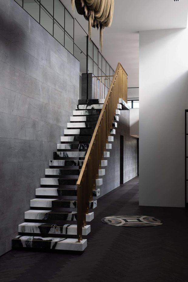 A dramatic contemporary interior focused on a sculptural open-riser staircase. The staircase has glossy black-and-white veined stone treads, a slender vertical timber balustrade, and a dark timber floor. Large grey stone-look wall panels, black-framed internal glazing, white walls, and a sculptural hanging light create a refined, high-contrast foyer or circulation space.