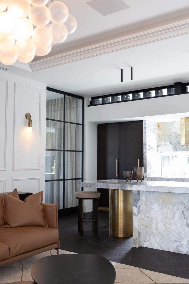 A refined open-plan living room and home bar with a contemporary classic interior. The space features white decorative wall panelling and ornate ceiling moulding, black-framed glazed partitions, dark timber flooring, a dramatic veined stone bar counter and splashback, a cylindrical brass-toned support, and a sculptural cluster pendant light. A tan leather armchair and dark round coffee table occupy the foreground.
