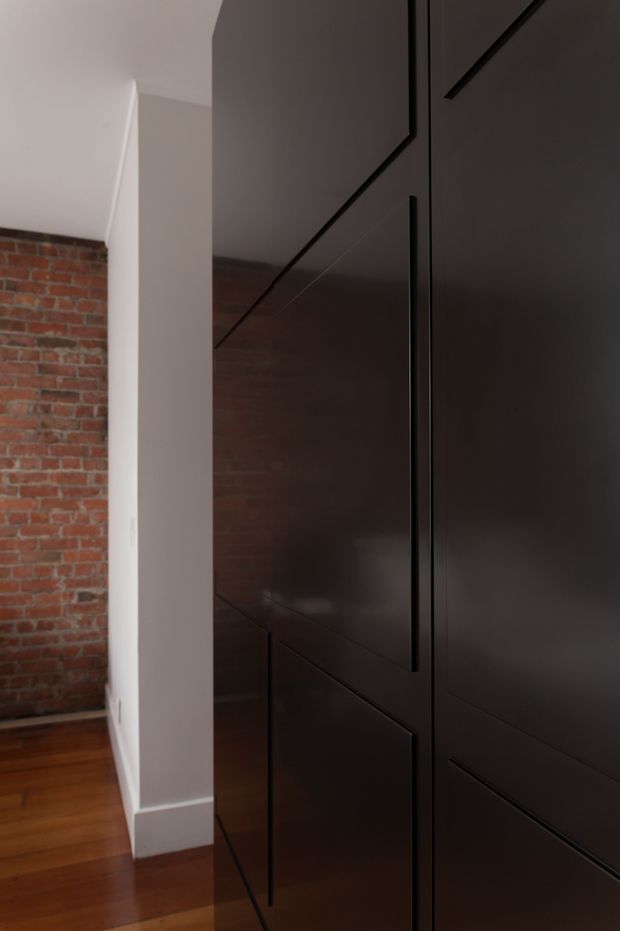 Close-up interior view of tall dark built-in cabinetry or paneled doors with geometric recessed detailing. A red exposed brick wall, white painted wall and trim, and polished timber flooring are visible beside the joinery, creating a refined industrial interior character.