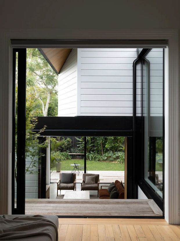View from a bedroom through a large black-framed glazed opening toward a covered outdoor sitting area and landscaped garden. The room has a warm timber floor and a low bed or upholstered seating in the foreground. The adjacent exterior structure features white horizontal weatherboard cladding, a timber-lined sloping soffit, and substantial dark structural framing around the glazing.