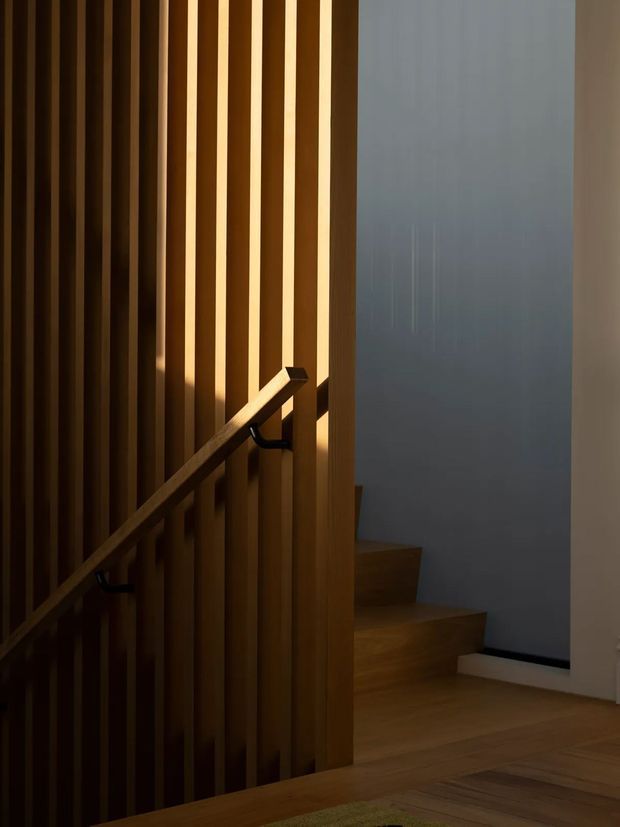 A moody interior view of a timber staircase and landing. Vertical timber slats form a balustrade or screening wall, with a simple matching timber handrail and warm concealed lighting between the slats. Timber stair treads and flooring continue toward a pale grey wall, creating a restrained contemporary interior.
