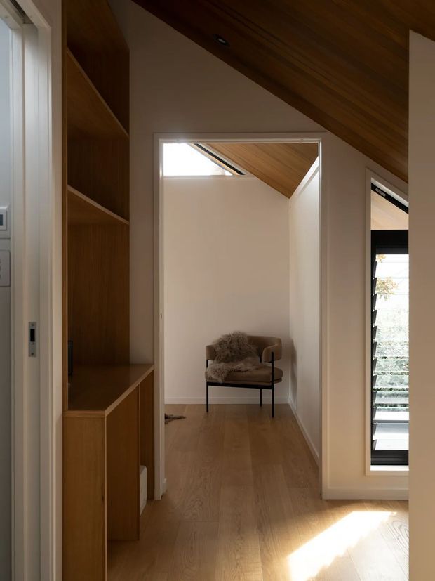A refined residential interior hallway or landing with warm timber flooring, integrated timber shelving and cabinetry, white walls, and a timber-lined sloping ceiling. The hallway leads to a small seating nook with a contemporary upholstered chair. A tall louvred window brings in strong daylight, while recessed lighting and carefully detailed joinery create a calm, minimalist atmosphere.
