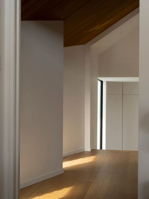 Minimalist interior hallway viewed through a doorway, with warm timber flooring and a sloped timber-clad ceiling. White plaster walls, simple white trims, and a tall narrow black-framed opening lead toward flush white built-in cabinetry. Natural light creates a bright, calm atmosphere.