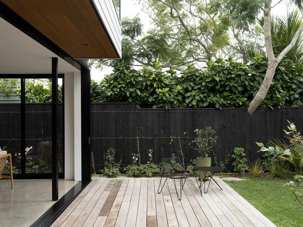 A contemporary residential outdoor deck and garden courtyard viewed from beside the house. The deck is clad in pale timber boards and furnished with a small outdoor table and chairs, while full-height black-framed glazing opens onto the space. A black timber boundary fence, dense hedging, mature trees and planted garden beds create a private, leafy setting. The house features a projecting timber-lined soffit and dark window framing.