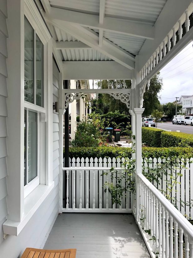 A narrow covered front porch or verandah on a white weatherboard house, featuring a grey-painted timber deck, white decorative timber balustrades and turned posts, ornate fretwork brackets, a corrugated roof with exposed painted rafters, and a white-framed window. A picket fence, clipped hedge, gardens, and residential street are visible beyond.