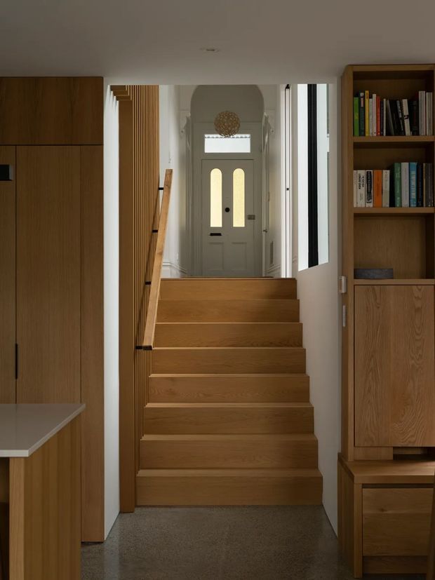 Interior view of a compact residential stairway leading up to a traditional-style entry hall. The staircase has warm timber treads and risers, a slim timber handrail, and vertical timber slat detailing along the side. Built-in timber cabinetry and open bookshelves frame the stairwell, while a pale front door with narrow arched glazed panels is visible at the landing. The lower floor appears to be a polished grey concrete or stone finish, and the space is softly lit with a warm, refined palette.
