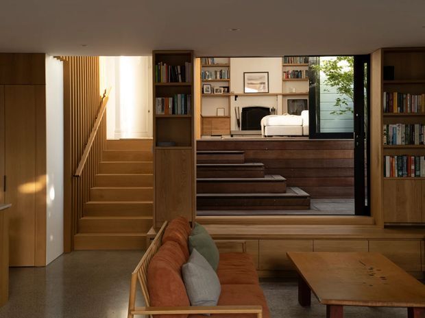 Warm, refined contemporary living area centered on a timber-framed opening to a raised sitting room. The foreground includes a low timber-framed sofa with rust and muted green cushions beside a substantial timber coffee table. Built-in timber cabinetry and open bookshelves frame the view, while broad timber steps lead upward past vertical timber screening to a higher-level room with a fireplace, bookshelves, artwork and glazing. The space has a layered split-level layout, subdued natural materials and soft afternoon light.