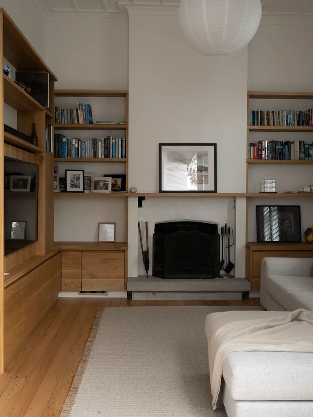 A warm, refined living room centred on a traditional fireplace with a black firebox and pale stone hearth. Custom timber built-in shelving and cabinetry flank the fireplace and continue along the walls, filled with books and framed photographs. A large neutral sectional sofa and textured area rug occupy the foreground, while a white paper pendant hangs from a detailed coffered ceiling. The room combines natural timber, cream walls, classic architectural trim, and restrained contemporary furnishings.
