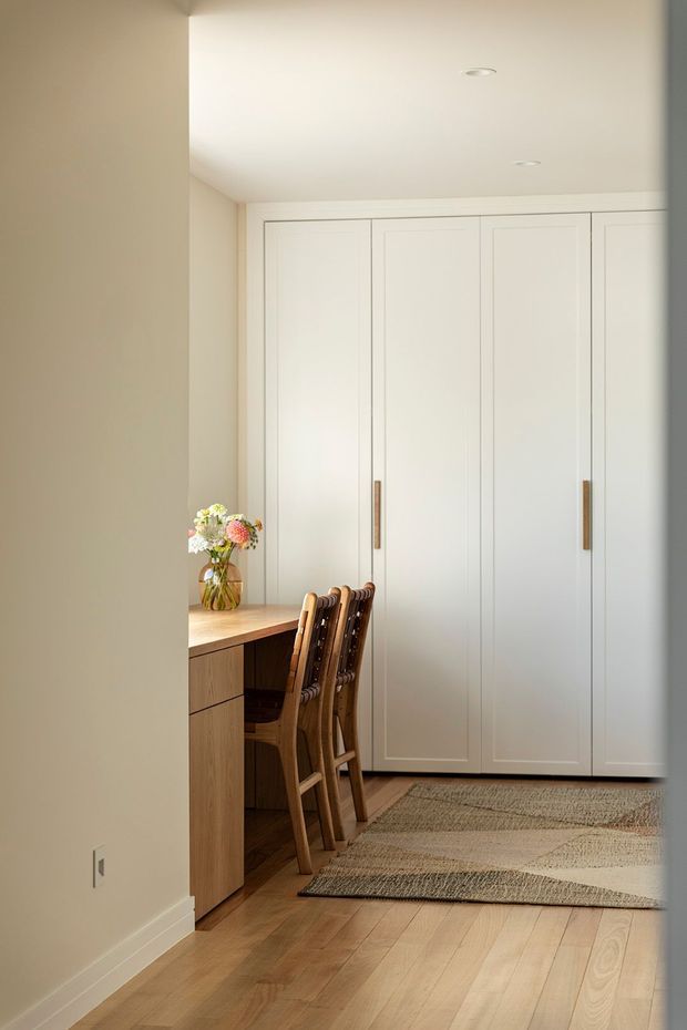 A warm, minimalist dining and built-in storage area with a pale timber dining table and several wooden chairs positioned along a wall. Full-height white paneled cabinetry with slim timber handles occupies the back wall, while light timber flooring and layered neutral woven rugs create a soft, natural palette. A small floral arrangement sits on the table, and recessed ceiling lights provide understated illumination.