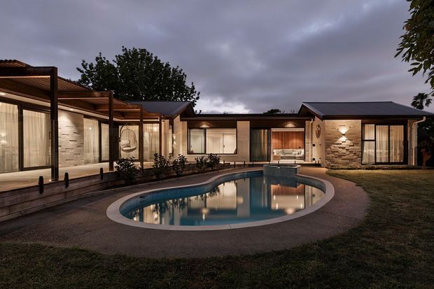 Twilight exterior view of a low-slung single-storey residence arranged around a curving swimming pool. The home features dark pitched roof sections, extensive floor-to-ceiling glazing, warm timber-lined covered outdoor areas, stone-clad feature walls, and illuminated interiors. The landscaped lawn and pool create a calm courtyard-like setting.