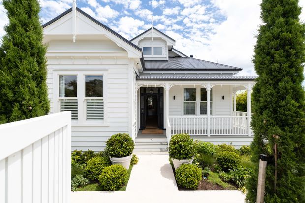 Front exterior view of a white weatherboard house with a gabled roof, dormer window, decorative verandah, white railings, and a landscaped entry path framed by tall narrow evergreen trees. The facade has a traditional heritage-inspired character with contrasting dark grey roofing and detailed timber trims.