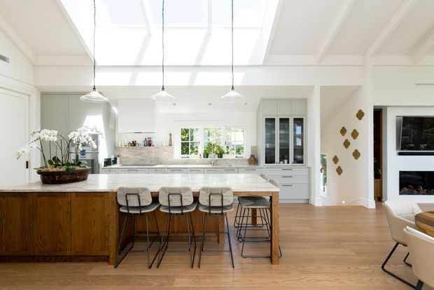 Bright, open-plan kitchen with a large timber island, four stools, white cabinetry, marble-look benchtops, pendant lights, a central skylight, and warm timber flooring. The room connects visually to a living area with a wall-mounted television and fireplace, while decorative wall objects and indoor plants add character.