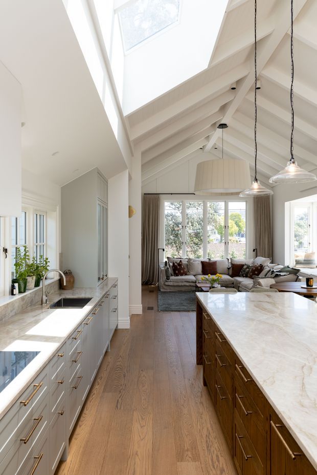 Bright open-plan kitchen and living area with a large marble island, pale built-in cabinetry, timber flooring, exposed white-painted rafters, and abundant natural light from a skylight and large windows. The kitchen opens directly toward a furnished sitting area with sofas and curtains.