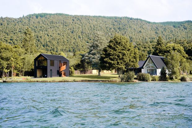 A wide exterior view across a body of water toward two small houses set in a landscaped, wooded site. The left house is a dark, modern two-storey structure with a steep gabled roof, large glazing and warm timber-toned elements. The right house is a compact white gabled building with dark trim and a covered entry. Forested hills form the background.