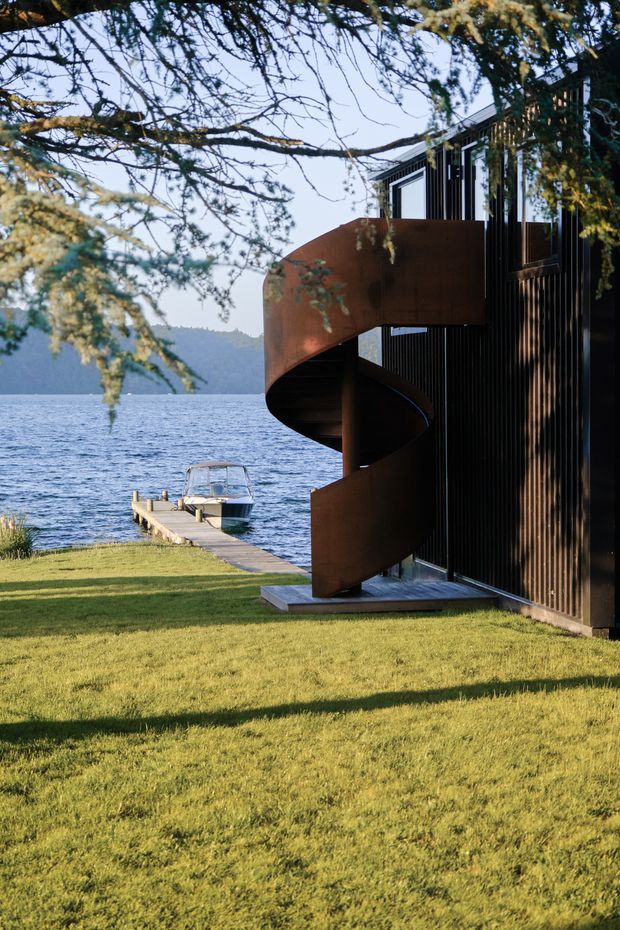 Exterior view of a contemporary lakeside building with dark vertical cladding and a prominent rusted steel spiral staircase wrapping around the facade. Floor-to-ceiling windows and a small dock with a boat sit beside the water, while mature trees frame the scene and a broad lawn extends in the foreground.