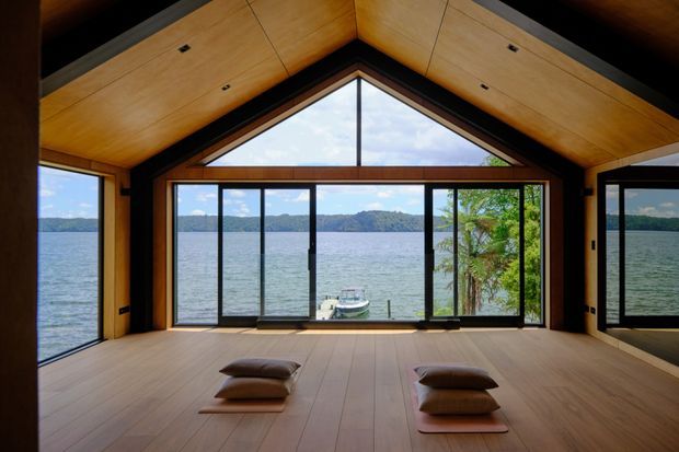 A serene timber-lined interior with a high gabled ceiling and expansive black-framed glazing opening directly onto a lake. The room has a warm wood floor and ceiling, minimal built-in detailing, and low floor cushions arranged in the foreground. The lake, boat, and tree-covered shoreline are visible beyond the glazing.