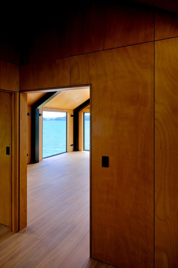 A dark, richly timber-lined interior corridor or room looking through an open doorway into a bright waterfront-facing space. Full-height warm wood wall panelling and matching timber flooring create a cohesive built-in interior, while the adjoining room has expansive glazing, a timber ceiling, and views across blue water and a distant shoreline. Black recessed hardware and dark structural trims provide contrast.