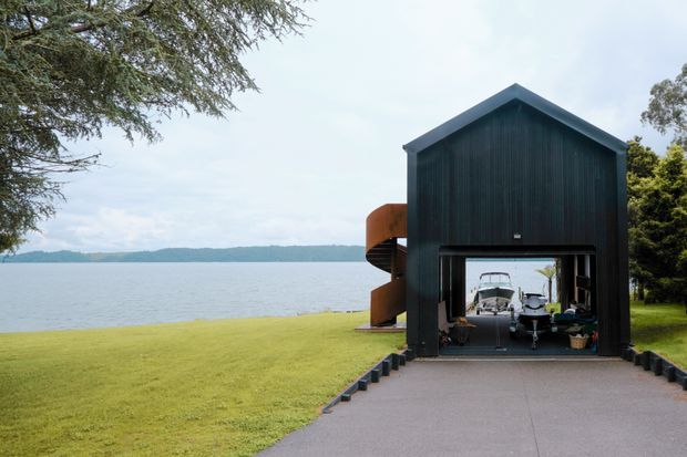 Exterior view of a dark, gable-roofed waterfront outbuilding or boathouse, clad in vertically ribbed dark material. A large open garage-style opening reveals boats and water beyond, with a curved orange-brown structure visible along the side. The building sits at the end of a paved drive beside a broad lawn and calm lake, with trees framing the scene.