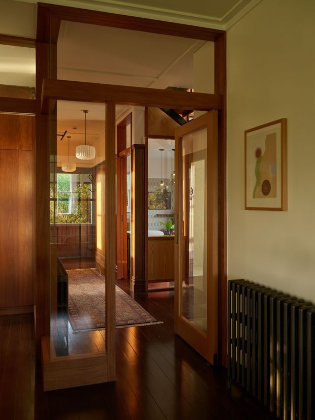 A warm, richly detailed interior view through a series of timber-framed glazed doors and partitions. The hallway features dark polished timber flooring, extensive dark wood joinery and trim, a patterned runner rug, pendant lights, framed artwork, and a black vertical radiator. Multiple connected spaces and leafy views are visible beyond the doorway, creating a layered, gallery-like circulation space.
