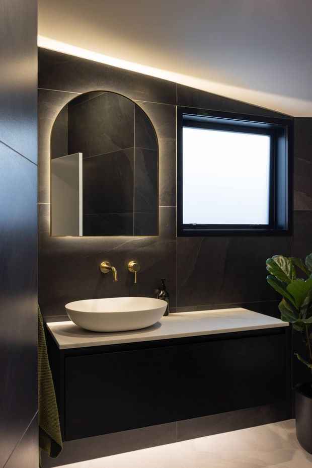 A moody contemporary bathroom vanity with large dark stone-look wall tiles, a floating black vanity, pale stone benchtop, white vessel basin, brushed brass wall-mounted tapware, and an arched backlit mirror. A black-framed window and concealed ambient lighting add contrast, while a plant and towel provide small natural accents.