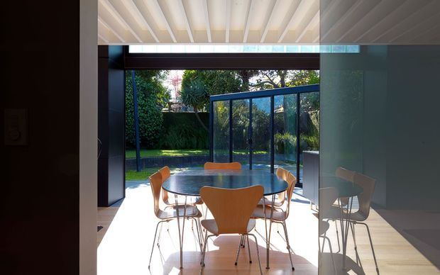 Bright dining area with a round dark dining table and sculptural wooden chairs, opening through extensive glazing to a landscaped garden and pool. The room has a white ribbed ceiling, pale timber flooring, dark wall framing, and a glazed corner enclosure that creates a strong indoor-outdoor connection. Reflections in the right-hand glass repeat the dining furniture.