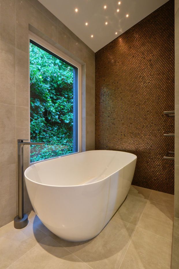 A contemporary bathroom featuring a freestanding white bathtub beside a large window overlooking dense greenery. Large beige stone-look tiles cover the walls and floor, while the main feature wall is clad in small bronze-toned mosaic tiles. Recessed ceiling lights and brushed metal bath fittings complete the refined, warm interior.