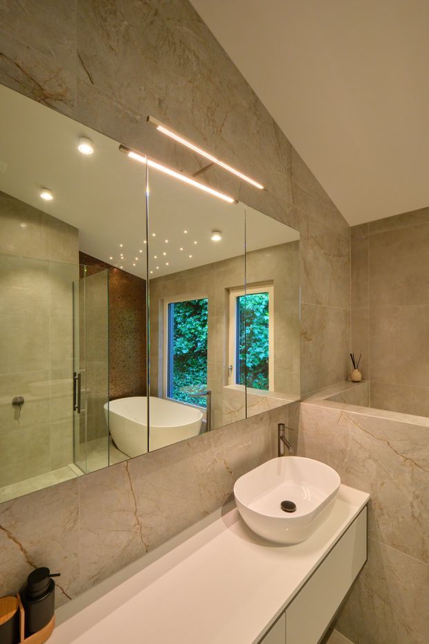 A refined contemporary bathroom clad in large-format beige and softly veined stone-look tiles. The room features a floating white vanity with an integrated countertop and white vessel basin, a freestanding bathtub, frameless glass shower enclosure, wall-mounted tapware, and a large mirrored wall. Linear LED lighting and recessed spotlights provide warm illumination, while narrow windows overlook greenery.